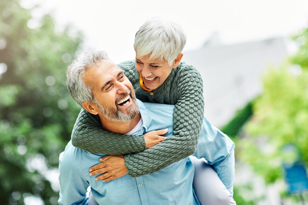 A smiling man giving a piggyback ride to a laughing woman outdoors.