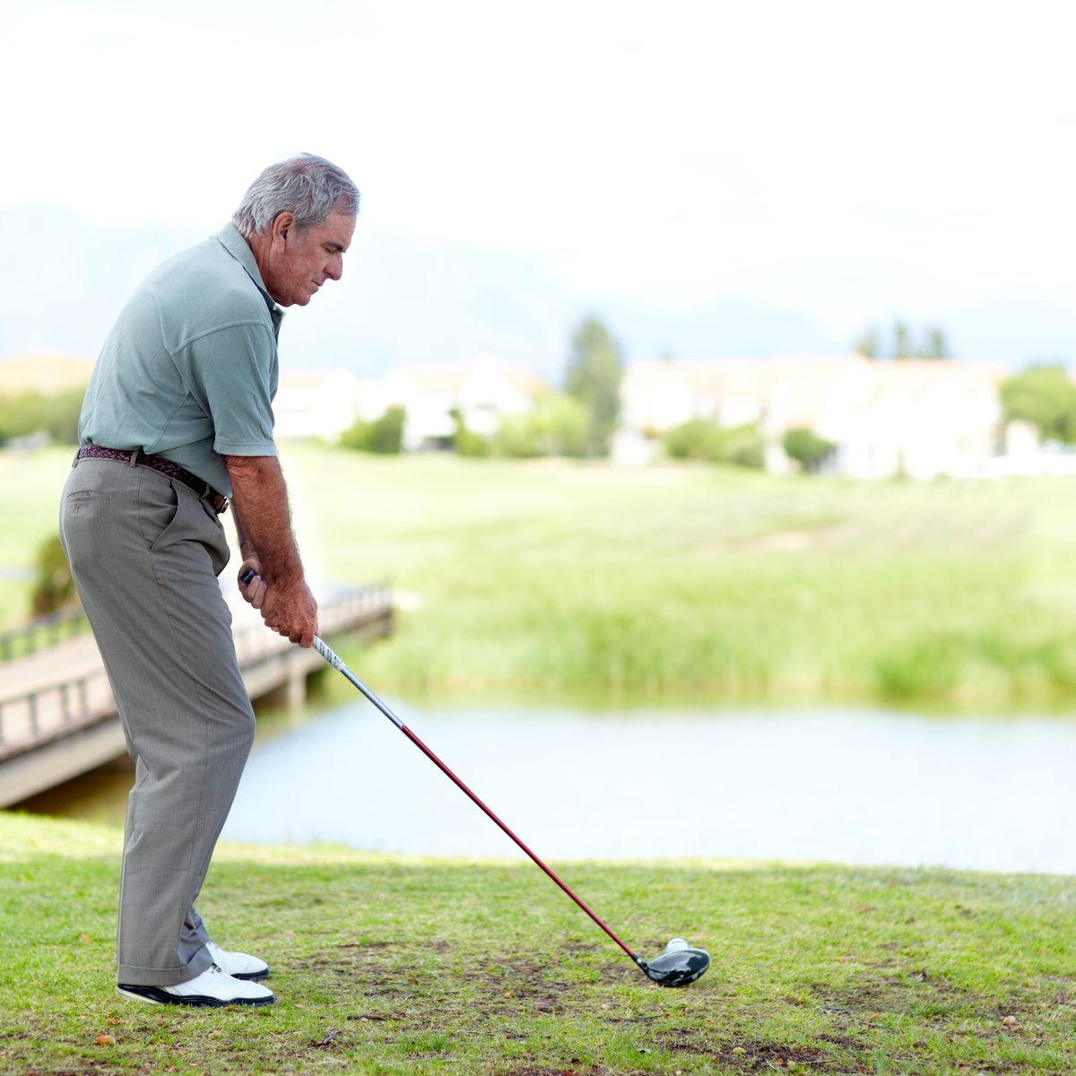 An older man in a golf shirt and slacks addresses a golf ball with a driver on a course.