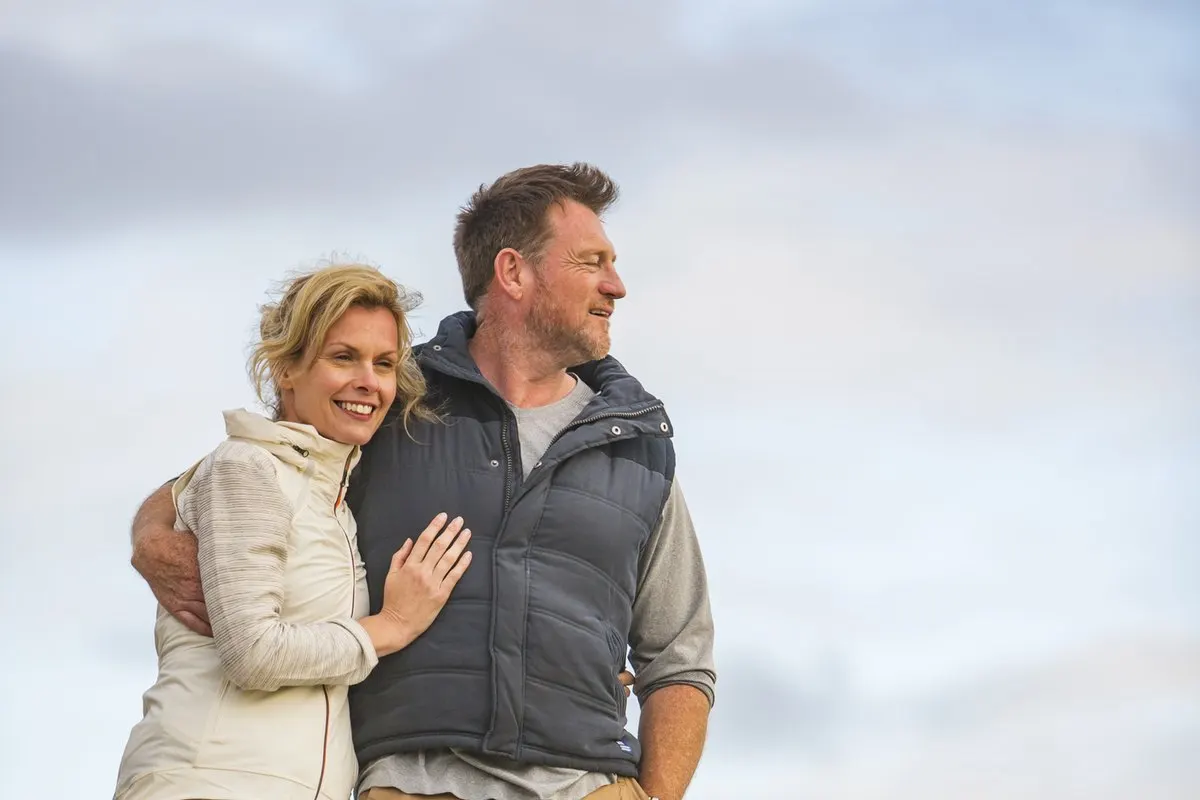 A man and woman standing close together and smiling while looking off into the distance.