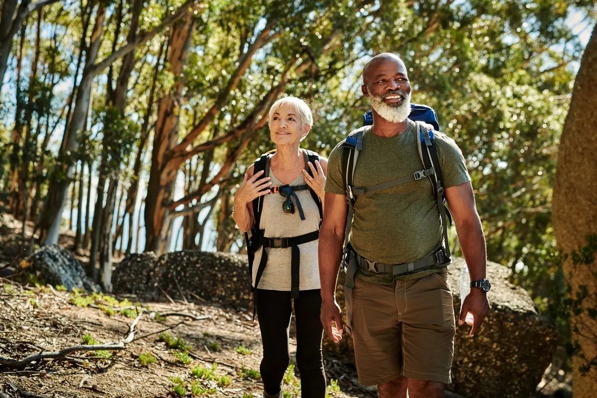 An older man and woman with backpacks hiking on a forest trail.