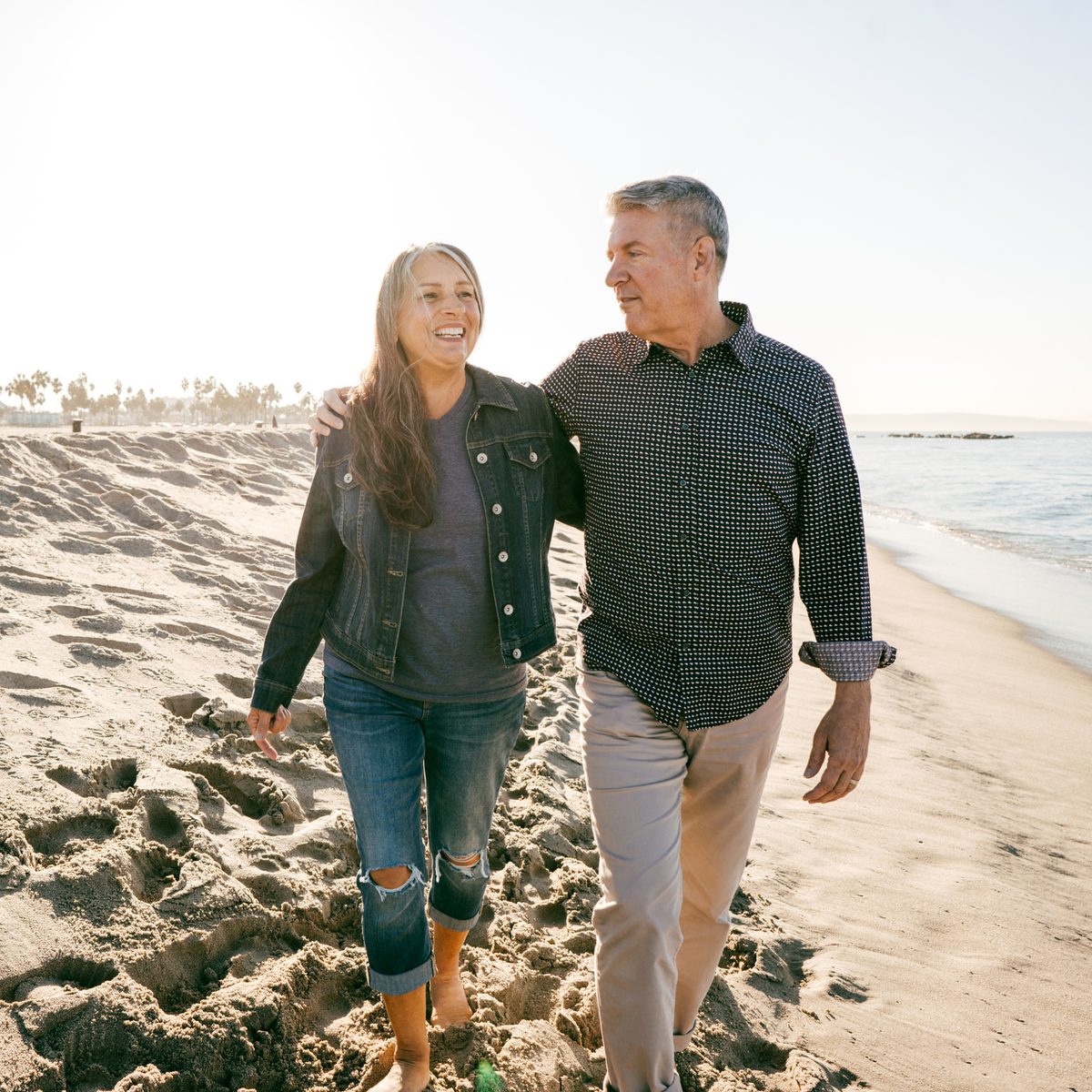 A middle-aged couple walks arm in arm along a sandy beach.