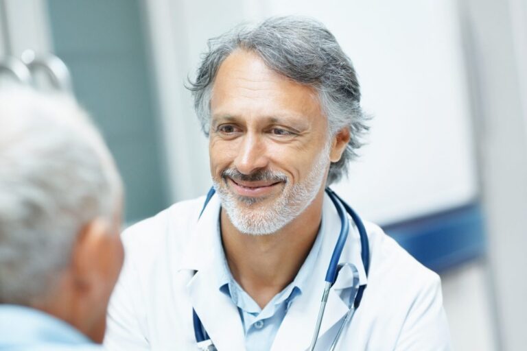 A smiling doctor with a stethoscope around his neck speaks with a patient.