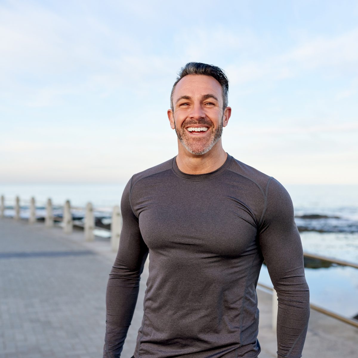 A smiling man in a long-sleeved gray athletic shirt standing on a paved walkway by the ocean.