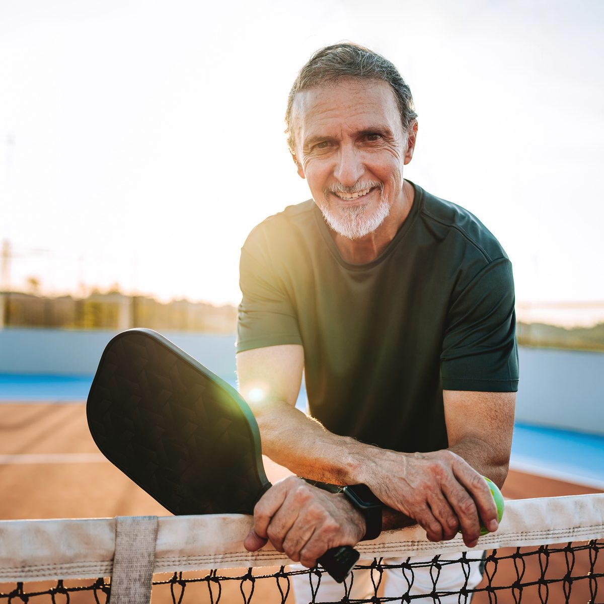 A smiling man leaning on a pickleball court net, holding a paddle and a ball.