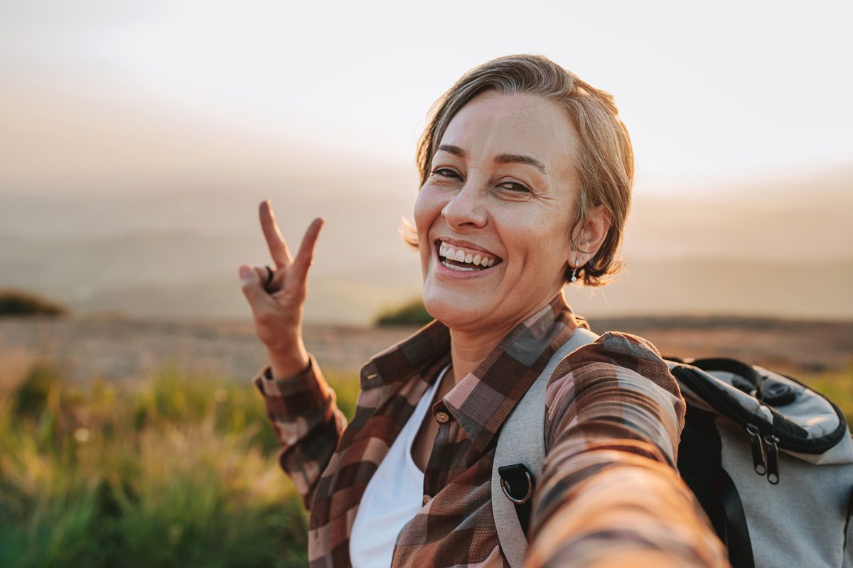 A smiling woman with a backpack takes a selfie while making a peace sign outdoors at sunset.