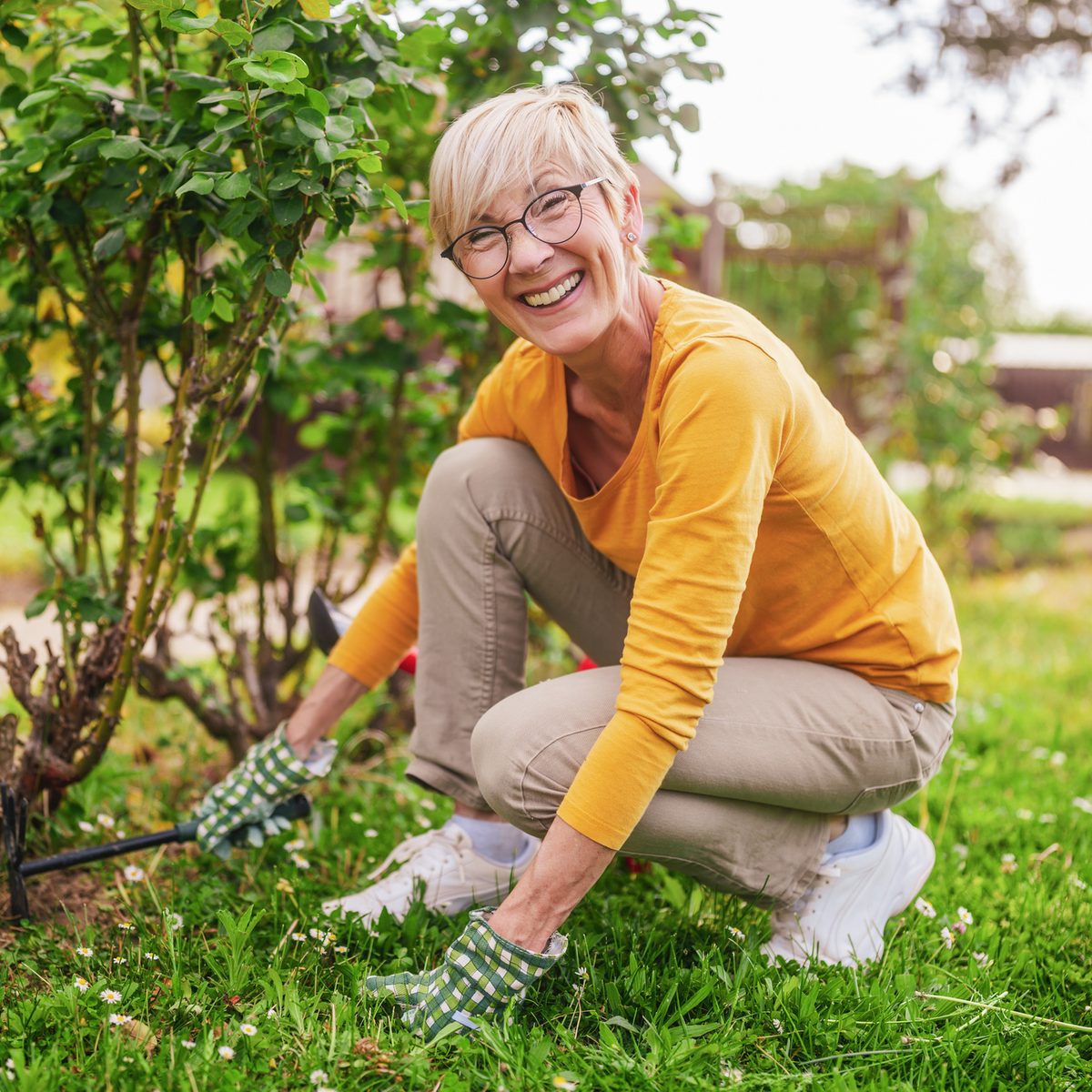 A smiling woman in a yellow long-sleeve shirt and gardening gloves crouches in a garden to prune a bush.