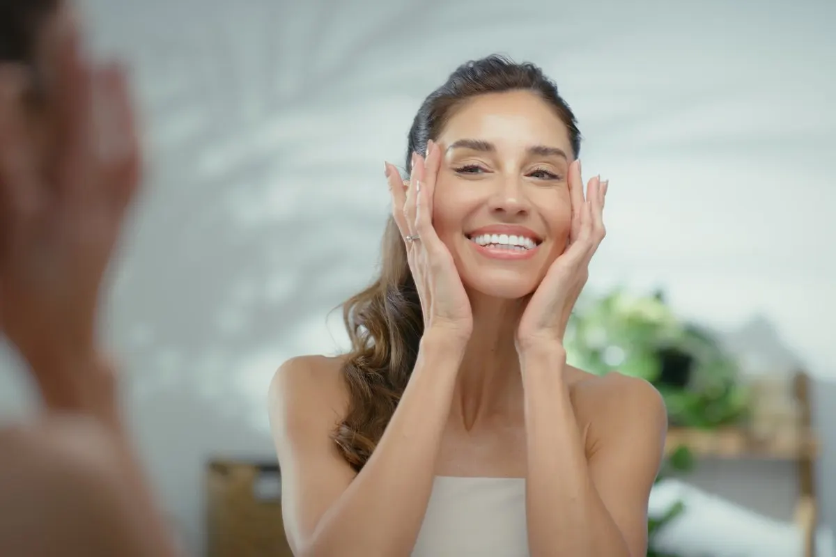 A smiling woman looking at her reflection in a mirror while gently touching her cheeks with both hands.