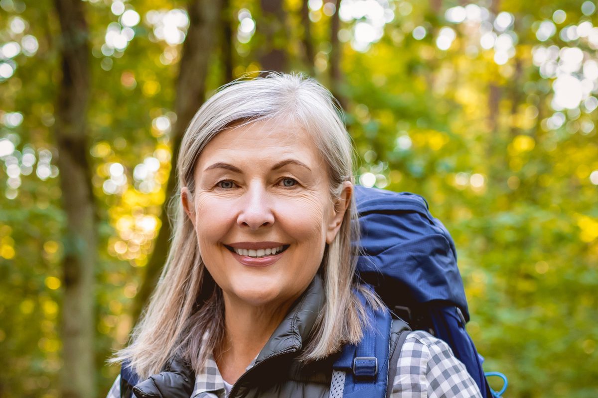 A smiling woman with long gray hair wearing a backpack in a forest.