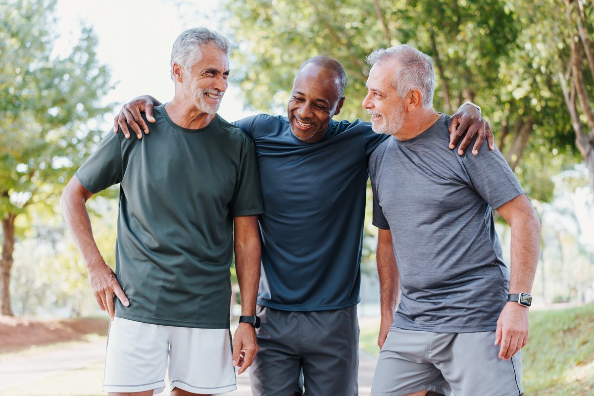 Three middle-aged men walking outdoors with their arms around each others shoulders, smiling and talking.