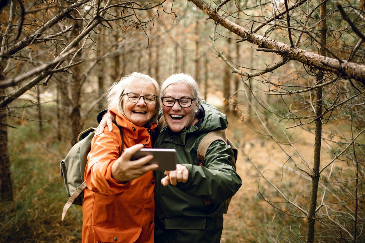 Two older women in hiking jackets smile and laugh while taking a selfie in a wooded area.