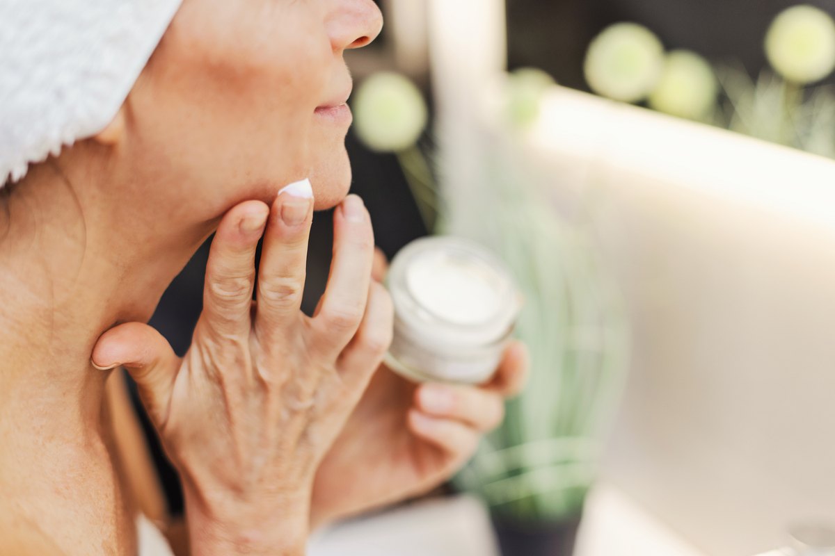 A person applying a dab of white face cream to their chin from an open glass jar.