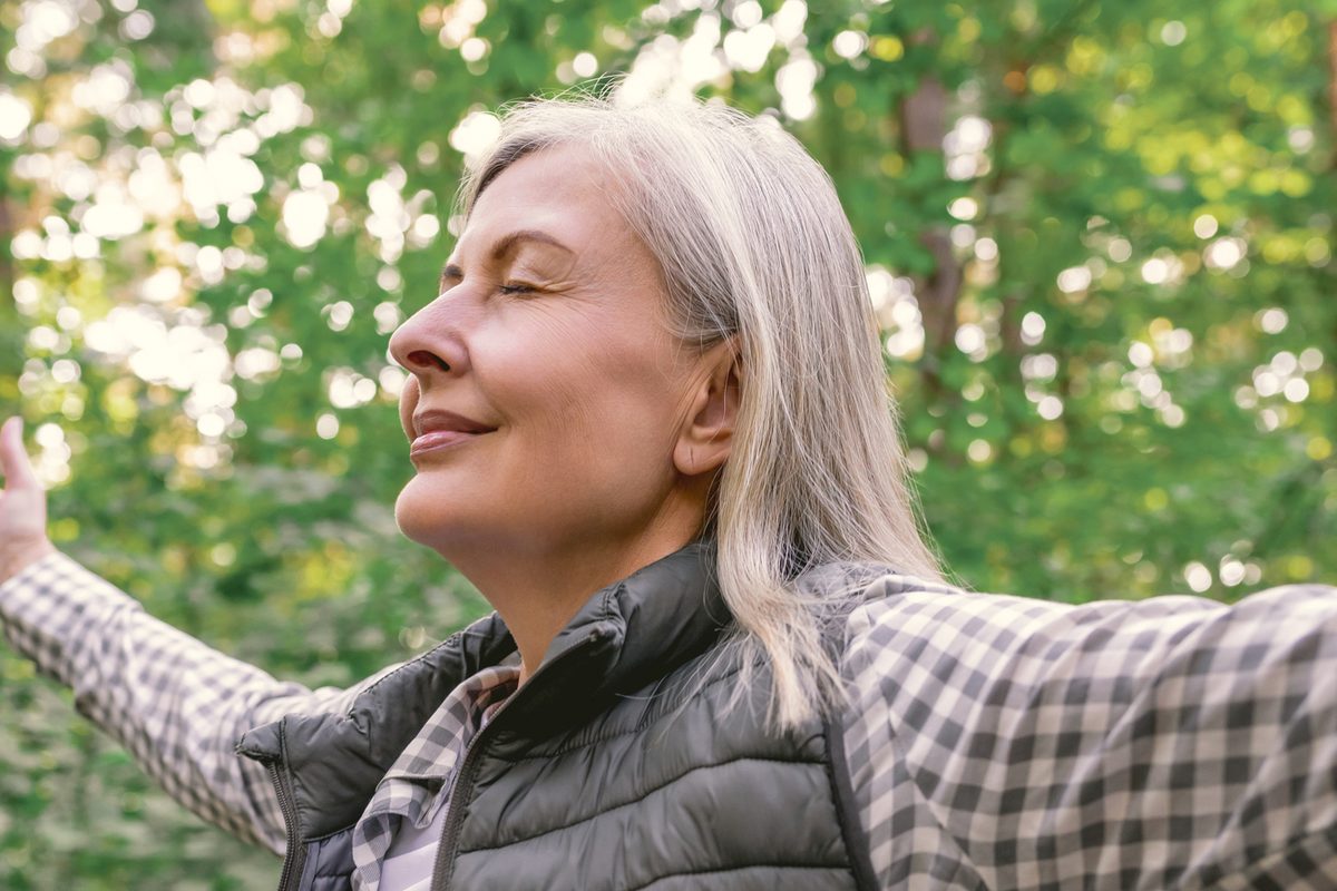 A middle-aged woman with grey hair standing outdoors with her eyes closed and arms outstretched.
